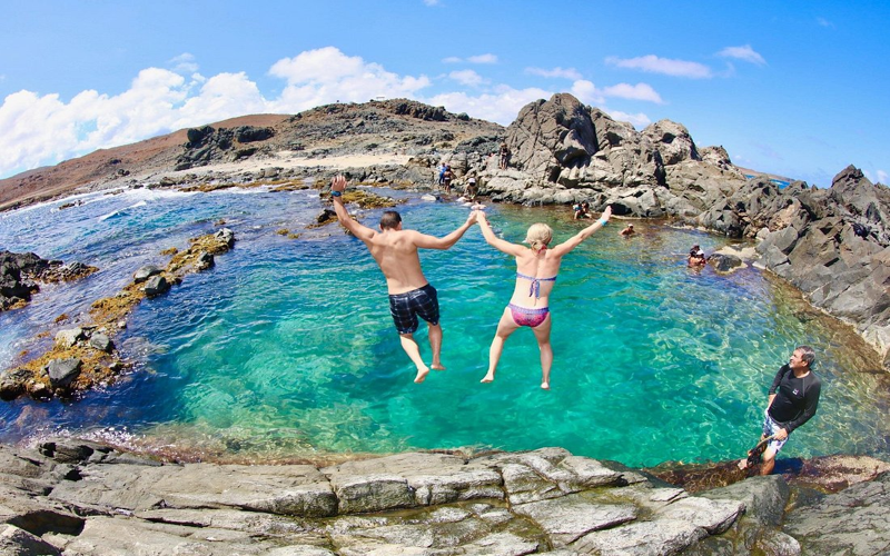 Two travelers jumping into the Conchi Natural Pool in Aruba while others swim and relax in the turquoise water.
