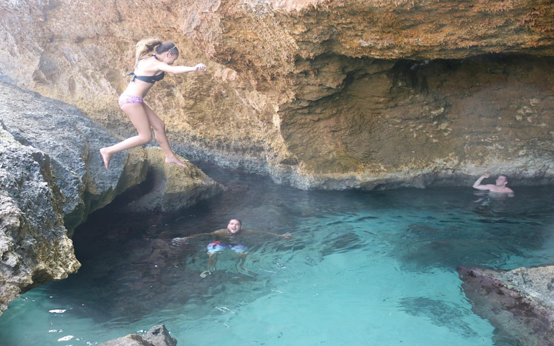 Traveler jumping into a hidden cave pool in Aruba while others swim and explore the natural rock formations.